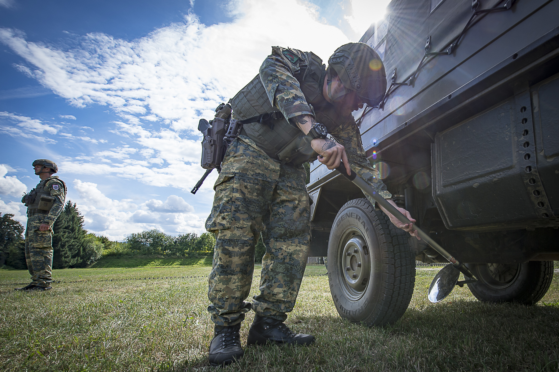 Ein Soldat untersucht mit Hilfe eines Spiegels die Unterseite eines Fahrzeuges.